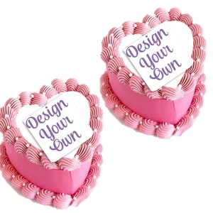 May include: Two pink heart-shaped cakes with white frosting and a scalloped edge. Each cake has a white square on top with the text "Design Your Own" in purple.