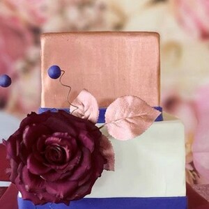 K&ouml;nnte beinhalten: Eine zweist&ouml;ckige Hochzeitstorte, verziert mit einer einzigen gro&szlig;en burgunderfarbenen Rose, einem einzelnen Blatt und blauen Akzenten. Die obere Etage ist kupferfarben, die untere Etage ist wei&szlig;, und beide Etagen haben ein blaues Band.