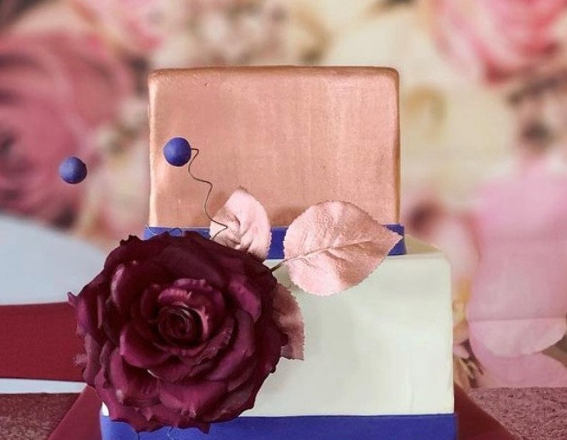 K&ouml;nnte beinhalten: Eine zweist&ouml;ckige Hochzeitstorte, verziert mit einer einzigen gro&szlig;en burgunderfarbenen Rose, einem einzelnen Blatt und blauen Akzenten. Die obere Etage ist kupferfarben, die untere Etage ist wei&szlig;, und beide Etagen haben ein blaues Band.