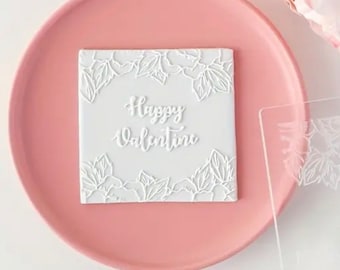 May include: A white square with the words "Happy Valentine" in cursive, surrounded by a floral design. The square is on a pink plate, with a clear plastic stencil nearby. A pink flower is in the background.
