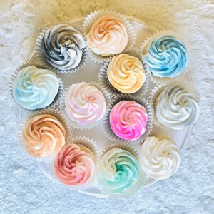 May include: A white cupcake stand with 12 cupcakes decorated with different colored frosting swirls. The cupcakes are arranged in a circle on the stand. The frosting colors include pink, blue, yellow, gray, and white.
