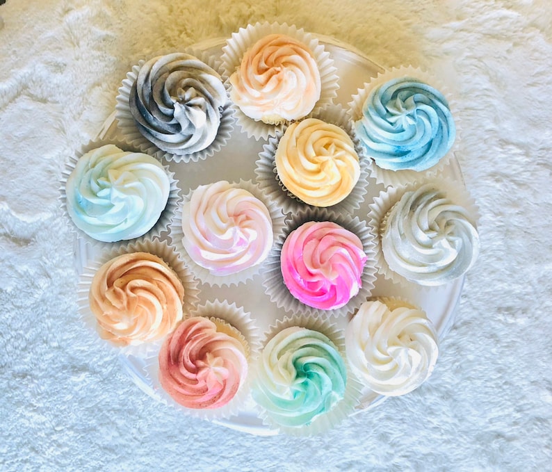 May include: A white cupcake stand with 12 cupcakes decorated with different colored frosting swirls. The cupcakes are arranged in a circle on the stand. The frosting colors include pink, blue, yellow, gray, and white.