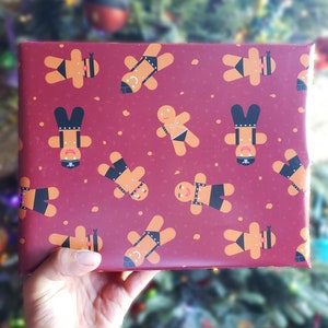 May include: Red wrapping paper with a repeating pattern of gingerbread people wearing black leather bondage gear. The gingerbread people are smiling and have white icing on their faces.