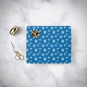 May include: Blue wrapping paper with a white snowflake pattern. A gold bow is on top of the paper.
