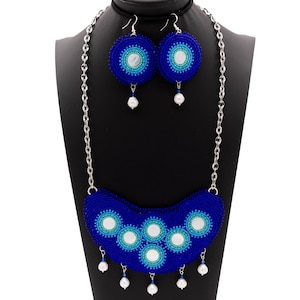 May include: A beaded jewelry set featuring a necklace and matching earrings. The necklace and earrings are primarily blue, with white and light blue accents. The necklace has a curved shape with multiple circular designs and dangling white pearls. The earrings are circular with a pearl drop.