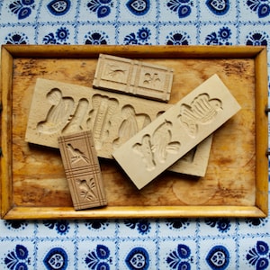 Könnte beinhalten: Ein Holztablett mit einer Auswahl an antiken Plätzchenausstechern. Die Ausstecher sind aus Holz gefertigt und zeigen verschiedene Designs, darunter Vögel, Blumen und geometrische Formen. Das Tablett ist aus hellem Holz gefertigt und hat eine natürliche Oberfläche.