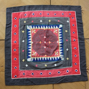 May include: A square, black fabric cushion with a red, white, blue, and yellow embroidered design. The center of the cushion features a circular pattern of spirals and geometric shapes.