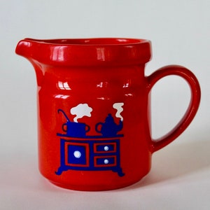 以下が含まれることがあります： 赤いセラミック製のクリーマーで、青と白のストーブのデザインが施されています。ストーブの上にはティーポットと鍋が置かれており、ストーブには白いノブが付いた引き出しが2つあります。