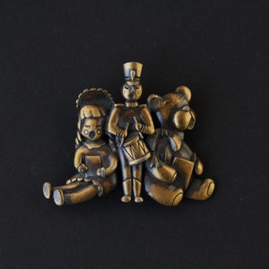 May include: A vintage gold-toned metal pin featuring a girl, a soldier, and a bear. The girl is holding a book, the soldier is playing a drum, and the bear is holding a book.