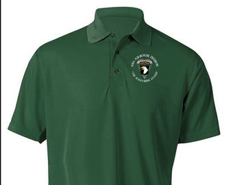 Może przedstawiać: Zielona koszulka polo z haftowanym logo 101. Dywizji Powietrznodesantowej na piersi. Logo przedstawia białego orła z parasolowcem i tekstem "101st Airborne Division The Screaming Eagles".