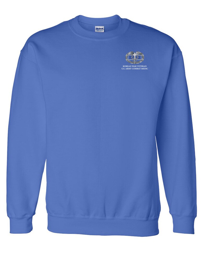 May include: A royal blue sweatshirt with a white embroidered patch on the left chest. The patch features a silver Korean War Veteran U.S. Army Combat Medic insignia.