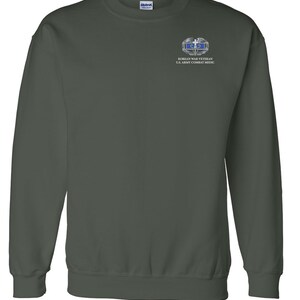 May include: A dark green sweatshirt with a small embroidered patch on the left chest. The patch features a silver and blue Korean War Veteran U.S. Army Combat Medic badge.