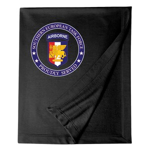 May include: Black fleece blanket with a white circle containing the text "Southern European Task Force", "Airborne", and "Proudly Served". The circle also features a blue and gold crest with a lion.