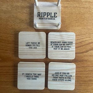 May include: A wooden coaster set with four square coasters and a holder. The coasters have inspirational quotes in black text. The holder is square with four wooden pegs. The text "RIPPLE HUNTER-GARCIA" is on the holder.