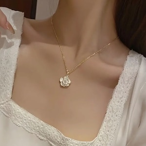 May include: A gold-colored necklace with a flower pendant. The pendant is a detailed, three-dimensional flower with a textured surface. The necklace is worn on a person, and the background is blurred.