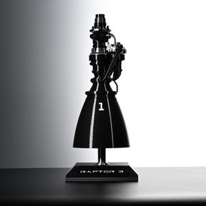 May include: A black Raptor 3 model rocket engine replica. The model features intricate details, a conical nozzle, and the number "1" in white. It stands on a black base with "RAPTOR 3" printed on it. A small red component rests nearby.