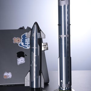 Op de afbeelding: Een zilveren model van de SpaceX Starship en Super Heavy raket. De Starship is een herbruikbaar ruimtevaartuig dat is ontworpen voor reizen naar de diepe ruimte, terwijl de Super Heavy een krachtige boosterraket is. De modellen worden getoond op zwarte bases met de namen "Starship" en "Super Heavy" erop gedrukt.