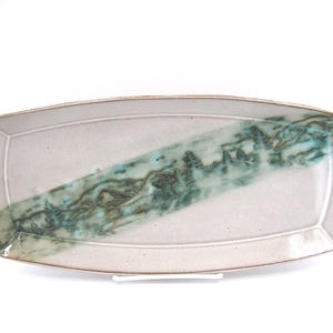 May include: A rectangular ceramic serving tray with a light beige, speckled base glaze. A diagonal band of textured, deep teal and forest green glaze runs across the center, featuring a subtle, abstract landscape pattern with dark brown accents.