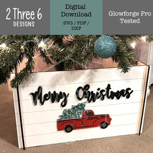 Op de afbeelding: Een witte houten doos met de woorden "Merry Christmas" in zwarte letters en een rode vrachtwagen met een groene kerstboom op de laadbak. De doos is versierd met een wit schuttingsontwerp.