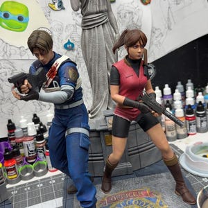 May include: Detailed figurines of video game characters, one in a blue uniform with a gun, the other in a red vest and shorts, also with a gun. A statue and the Raccoon Police Department logo are also visible.