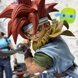 May include: A detailed figurine of an anime character with spiky red hair, green eyes, and a determined expression. The figure is dressed in a blue and green outfit, holding a sword. The figure has a white headband and a yellow scarf.