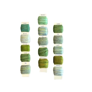 May include: A selection of green and blue yarn spools, labelled with fibre types: Cotton, Bamboo, Silk, and Silk/Linen. The spools are arranged in vertical rows, displaying a range of shades from light to dark green.