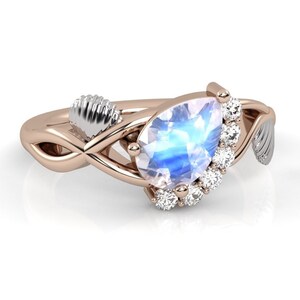 May include: A rose gold ring featuring a pear-shaped moonstone set in a halo of diamonds. The ring has a delicate, intertwined band with a small, white metal leaf on one side.