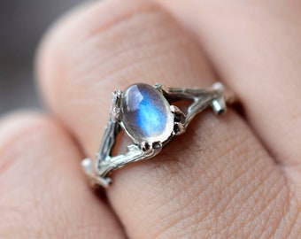 Puede incluir: Un anillo de plata con una piedra de luna azul engastada en un diseño de rama.