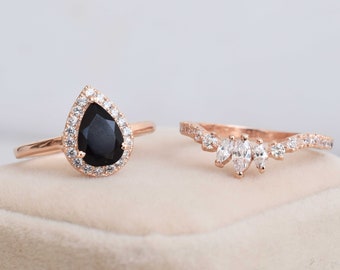May include: Two rose gold rings with clear and black gemstones. The larger ring features a pear-shaped black gemstone surrounded by a halo of smaller clear gemstones. The smaller ring has a curved band with five marquise-shaped clear gemstones.