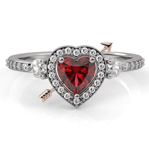 Könnte beinhalten: Ein silberner Ring mit einem roten herzförmigen Edelstein, der von einem Heiligenschein aus kleineren weißen Edelsteinen umgeben ist. Ein Pfeil mit einer goldenen Spitze durchbohrt das Herz und symbolisiert die Liebe.