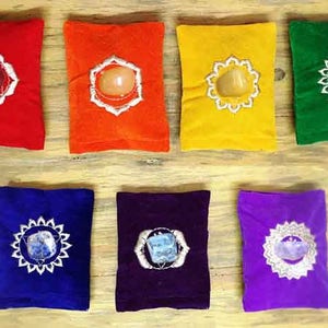 Könnte beinhalten: Sieben farbige Chakra-Kissen in Rot, Orange, Gelb, Grün, Blau, Indigo und Lila. Jedes Kissen hat einen Edelstein in der Mitte, umgeben von einem weißen Stickmuster. Die Kissen sind auf einer hellen Holzoberfläche angeordnet.