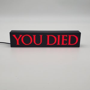 May include: A rectangular black light-up sign with the words "YOU DIED" in bright red letters. The sign is powered by a black cord and sits on a white surface. The sign is a decorative item.