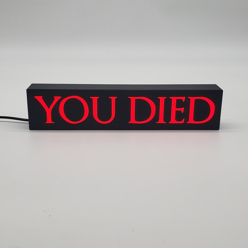 May include: A rectangular black light-up sign with the words "YOU DIED" in bright red letters. The sign is powered by a black cord and sits on a white surface. The sign is a decorative item.