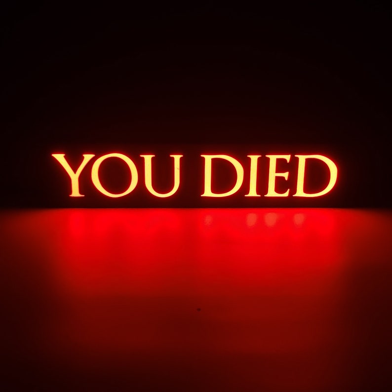 May include: Illuminated sign with the words "YOU DIED" in a bold, yellow font. The text is set against a dark background with a vibrant red glow emanating from below, creating a dramatic effect.