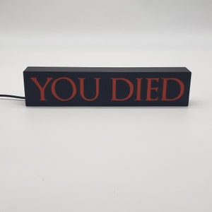 May include: A rectangular black sign with the words "YOU DIED" in red lettering. The sign is illuminated and has a power cord attached. The sign is likely a decorative item or a gaming-related product.