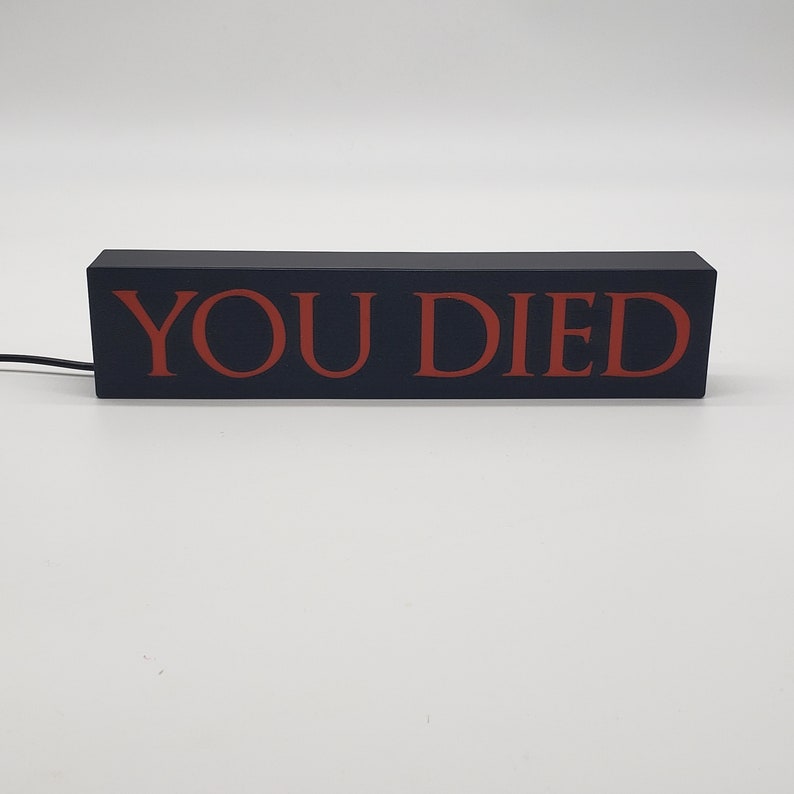 May include: A rectangular black sign with the words "YOU DIED" in red lettering. The sign is illuminated and has a power cord attached. The sign is likely a decorative item or a gaming-related product.