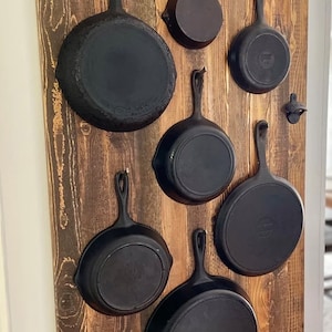 Peut inclure: Un porte-ustensiles mural en bois rustique avec plusieurs poêles en fonte accrochées à des crochets. Les poêles sont de différentes tailles et ont une finition noire.