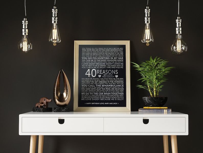 May include: A black and white printable poster with a white background and black text. The text lists 40 reasons to love a dad. The poster is framed in a light wood frame and is sitting on a white desk with a gold metal sculpture and a potted plant.