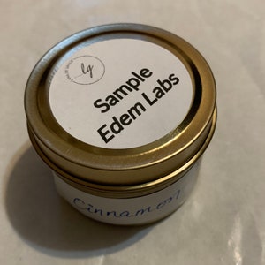 Puede incluir: Un pequeño tarro de velas redondo con tapa dorada. La etiqueta dice "Sample Edem Labs" y "Cinnamon" está escrito a mano con tinta azul en el tarro. El tarro está sobre una superficie blanca.