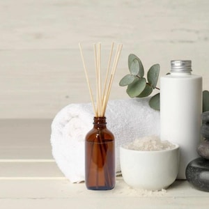 Könnte beinhalten: Ein Spa-Stillleben mit einer braunen Glas-Diffusorflasche mit Schilf, einem weißen Handtuch, einer weißen Keramikschale mit Salz, einer weißen Flasche und schwarzen Steinen. Eukalyptusblätter sorgen für einen Hauch von Grün.
