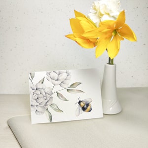 Pode incluir: Um cart&atilde;o de felicita&ccedil;&otilde;es em aquarela com fundo branco que apresenta um desenho floral com flores brancas e folhas verdes. Um zang&atilde;o est&aacute; voando perto das flores. O cart&atilde;o est&aacute; sobre uma superf&iacute;cie bege claro.