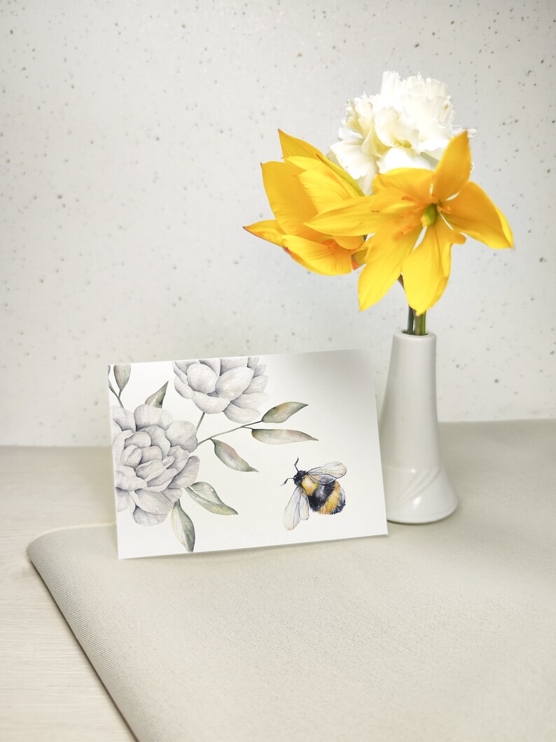 Pode incluir: Um cart&atilde;o de felicita&ccedil;&otilde;es em aquarela com fundo branco que apresenta um desenho floral com flores brancas e folhas verdes. Um zang&atilde;o est&aacute; voando perto das flores. O cart&atilde;o est&aacute; sobre uma superf&iacute;cie bege claro.