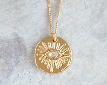 May include: Gold necklace with a round pendant featuring a stylised eye with a small clear stone in the centre. The pendant is surrounded by radiating lines.