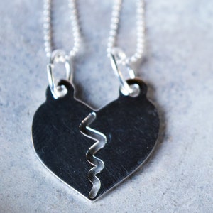 Può includere: Due collane d'argento con un ciondolo a forma di cuore spezzato. Il ciondolo è composto da due metà che si incastrano. Le collane sono su uno sfondo grigio con fiori di lavanda.