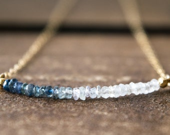 May include: A gold chain necklace with a graduated row of blue and white gemstone beads. The beads are arranged in a gradient from dark blue to light blue to white.