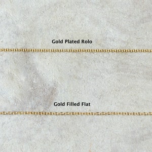 Pode incluir: Duas correntes de ouro sobre uma superf&iacute;cie cinzenta texturizada. A corrente superior &eacute; etiquetada como "Gold Plated Rolo" e a corrente inferior &eacute; etiquetada como "Gold Filled Flat".