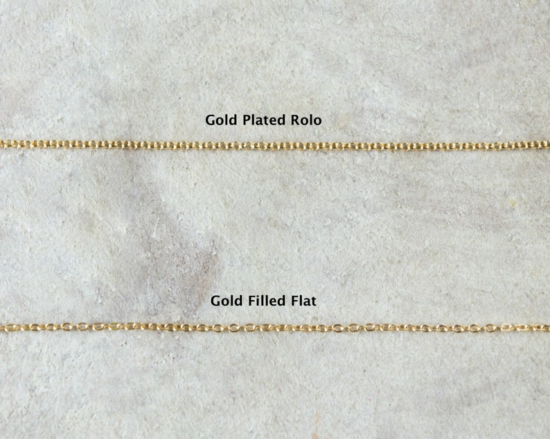 Pode incluir: Duas correntes de ouro sobre uma superf&iacute;cie cinzenta texturizada. A corrente superior &eacute; etiquetada como "Gold Plated Rolo" e a corrente inferior &eacute; etiquetada como "Gold Filled Flat".
