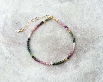 May include: A dainty bracelet featuring small, colourful gemstone beads. The beads are arranged in a gradient pattern, starting with pink and transitioning to green. A small gold cross charm hangs from the chain.