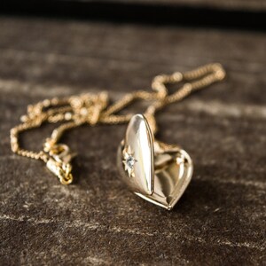 Pode incluir: Um colar de ouro com um pingente em forma de cora&ccedil;&atilde;o, adornado com uma pequena pedra que parece um diamante na parte da frente. O pingente est&aacute; aberto, revelando o interior.