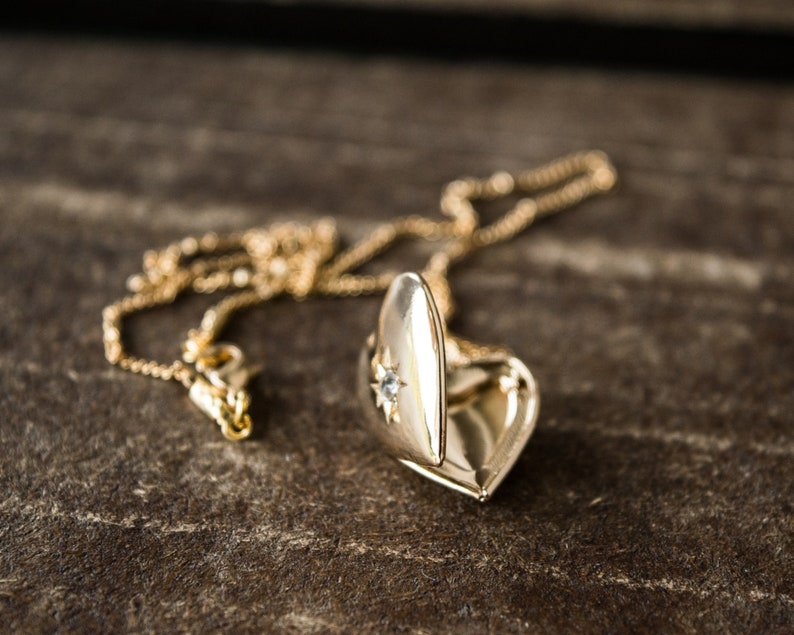 Pode incluir: Um colar de ouro com um pingente em forma de cora&ccedil;&atilde;o, adornado com uma pequena pedra que parece um diamante na parte da frente. O pingente est&aacute; aberto, revelando o interior.